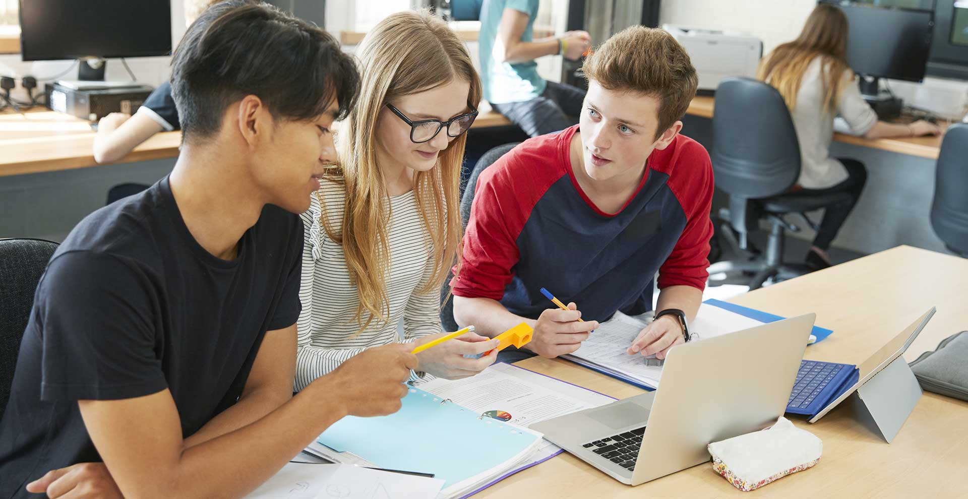 Drei jugednliche Schüler und Schülerinnen sitzen gemeinsam in einem Klassenzimmer vor einem Laptop und diskutieren.