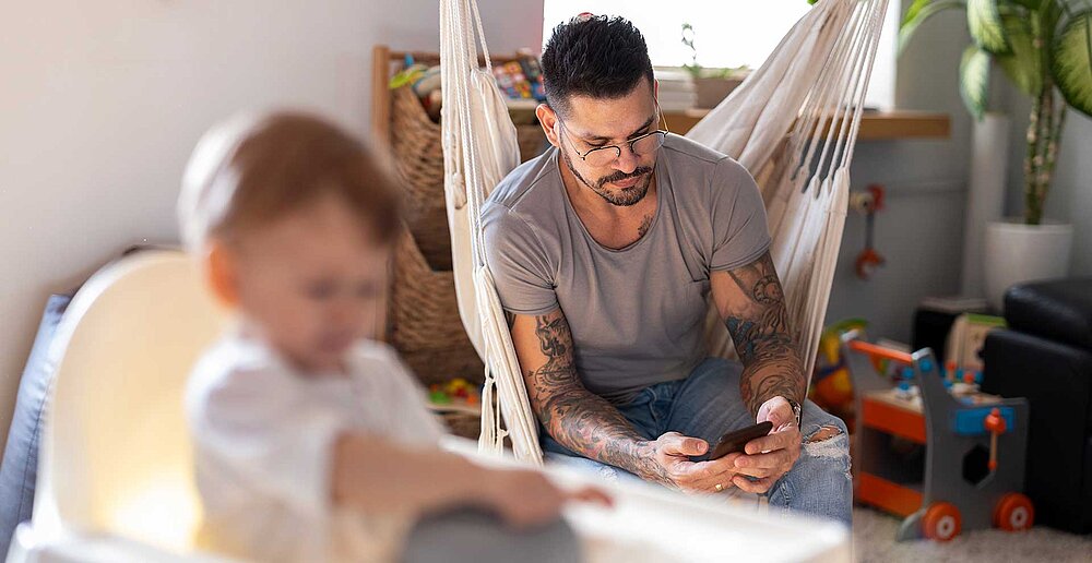 Junger Vater schaut auf sein Handy, das Kind sitzt im Hochstuhl