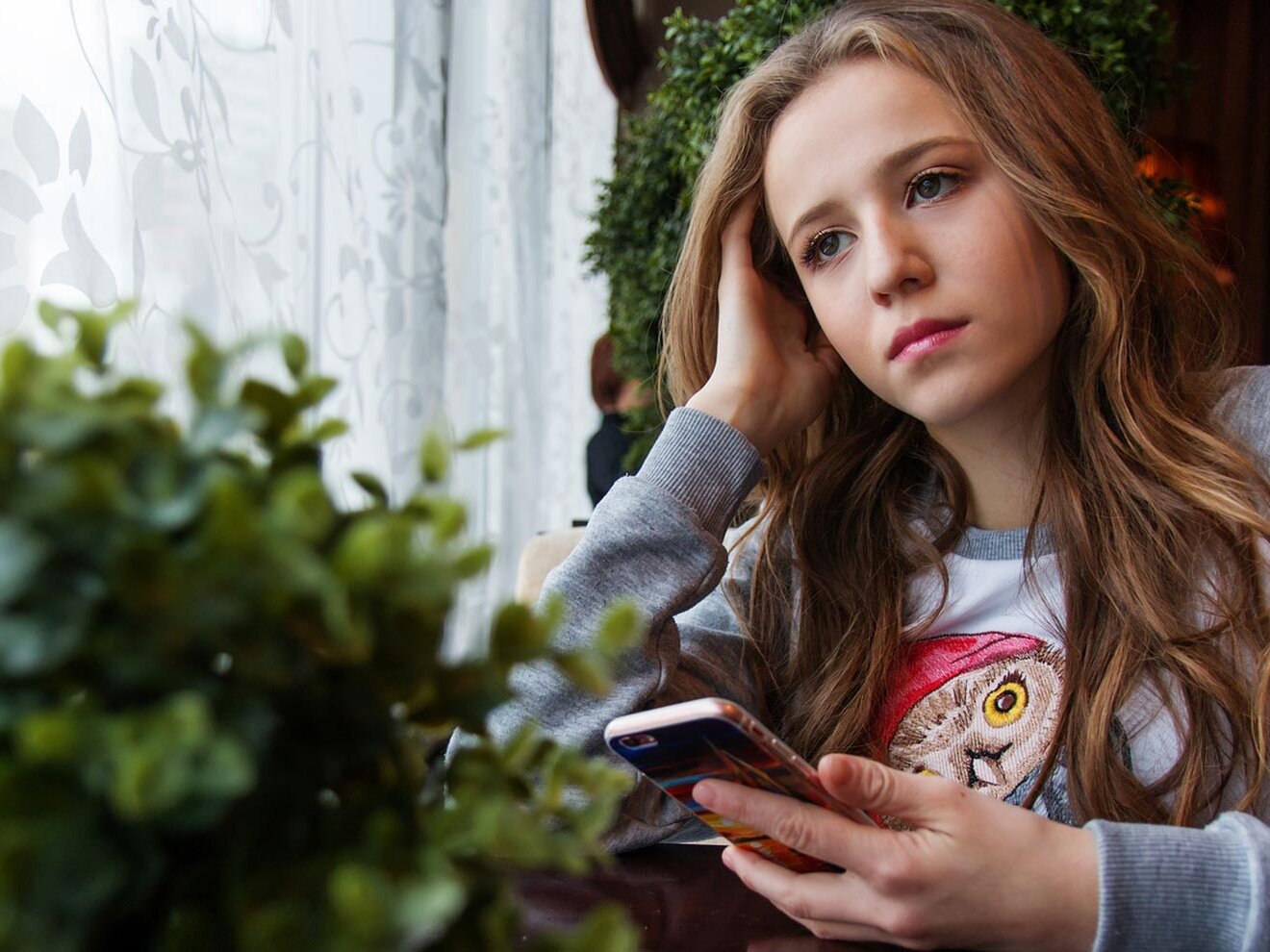 Mädchen mit Smartphone in der Hand schaut traurig aus dem Fenster