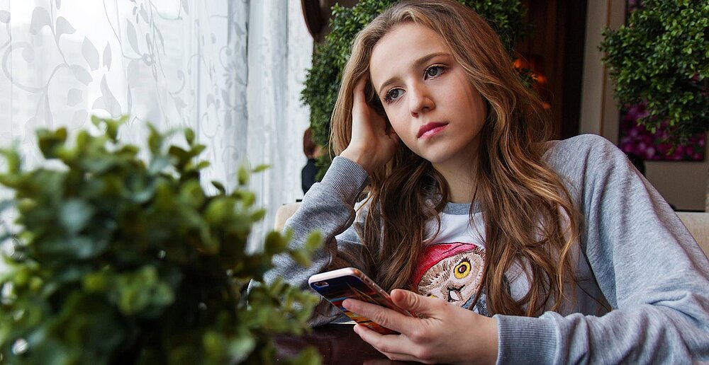 Mädchen mit Smartphone in der Hand schaut traurig aus dem Fenster