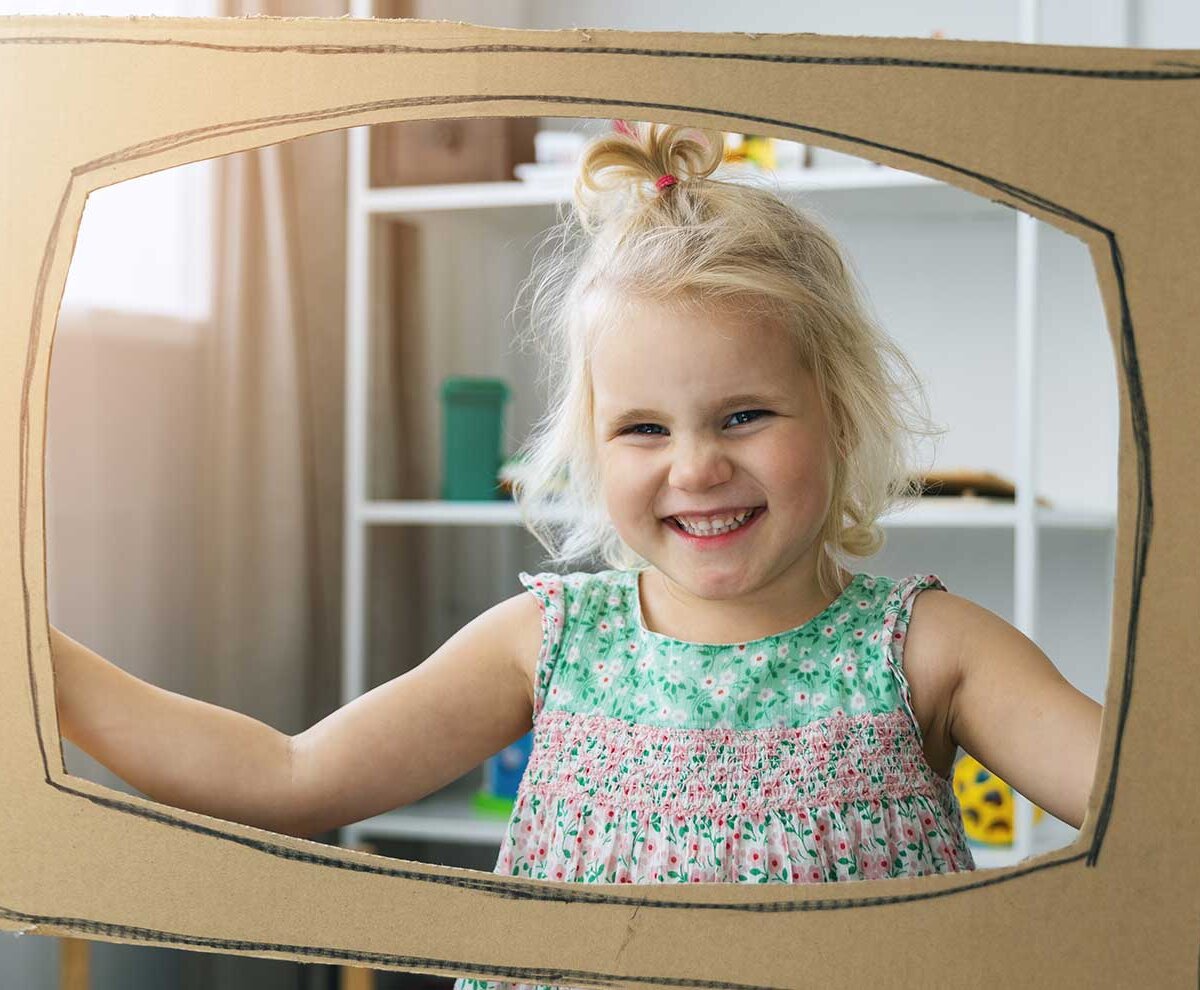 Ein kleines Mädchen schaut durch eine Pappschablone in Form eines Fernsehers.