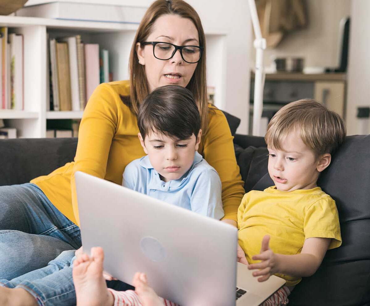 Mutter sitzt mit ihren beiden Kindern auf dem Sofa und gibt Anweisungen am Laptop.