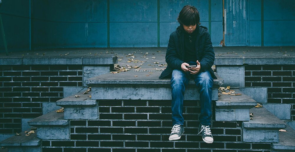 Junge mit Smartphone in der Hand sitzt traurig auf einer Treppe
