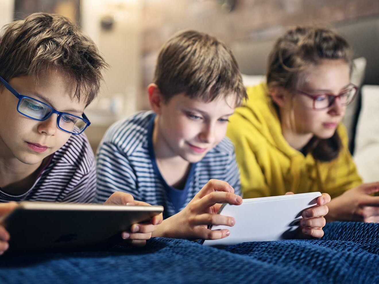 Drei Jugendliche liegen auf einem Bett und schauen auf verschiedene elektronische Geräte.